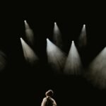 A solo performer stands in a spotlight on a dark stage during a dramatic moment.